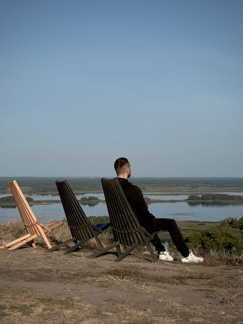 Free A man sits on a chair overlooking a peaceful river landscape on a summer day. Stock Photo