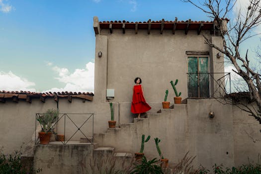 A woman in a vibrant red dress stands on a rustic staircase outdoors, against a clear blue sky.