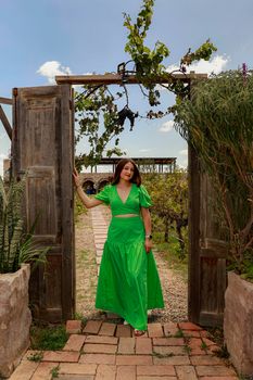 Woman in a vibrant green dress standing at a rustic wooden garden entrance in a vineyard setting.