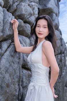 A young woman in a white dress standing gracefully beside a rocky wall outdoors.
