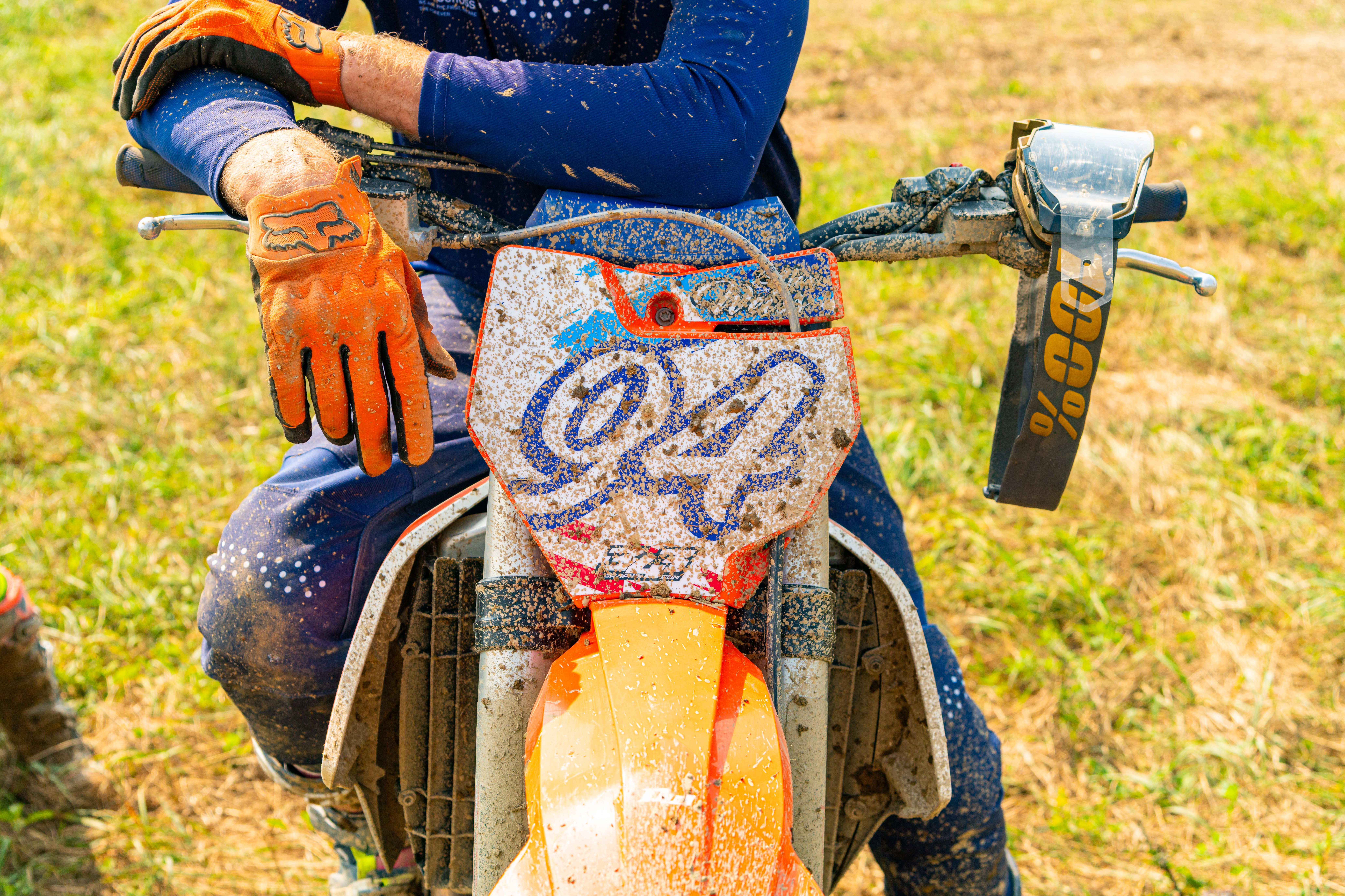 Motocross rider resting on a dirt bike, covered in mud during a sunny race day in Pennsylvania.