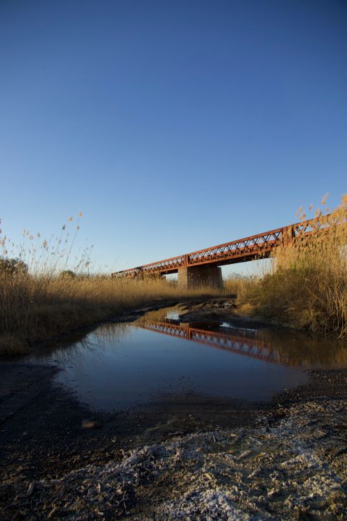 Free A serene view of a rustic metal bridge reflecting in a tranquil pool at sunset. Stock Photo