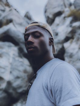 Young black man in a cap and t-shirt standing in front of rocky background, outdoors in Abuja, Nigeria.