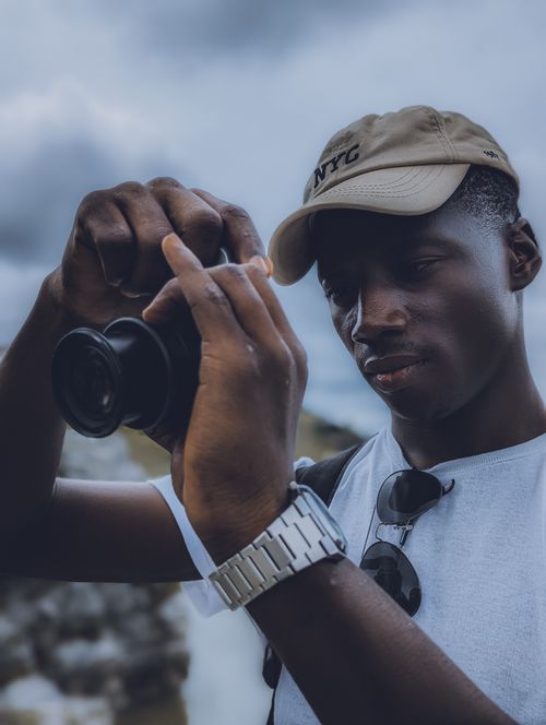 Free Young man in casual attire adjusting camera lens outdoors under cloudy sky. Stock Photo