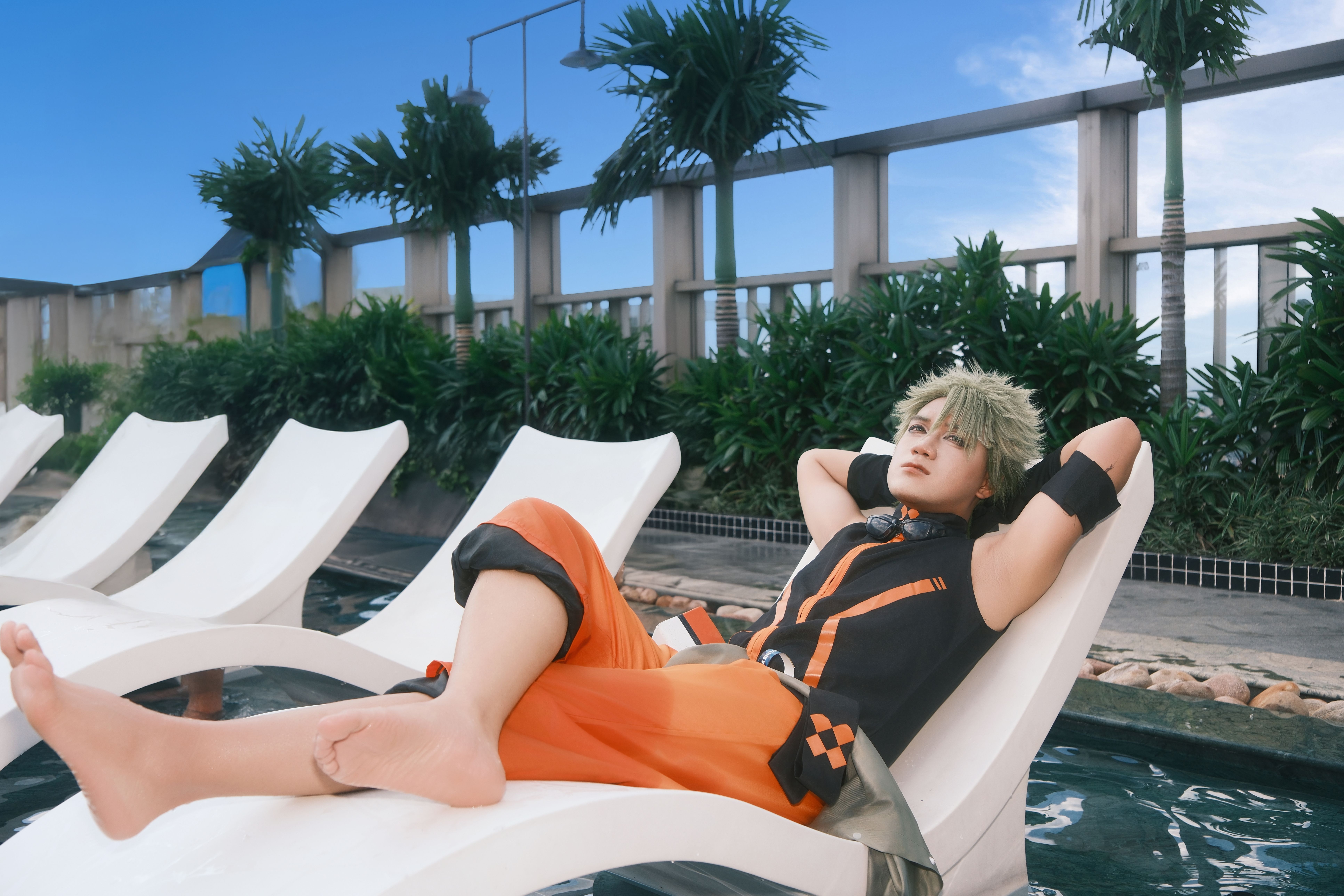 A person in vibrant cosplay attire relaxing on a poolside lounger, set against a lush, tropical backdrop with a clear blue sky.