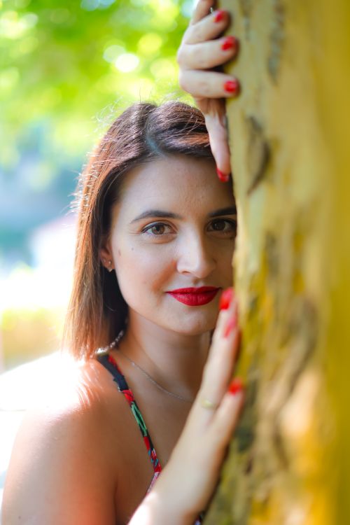 Free A woman with red lipstick poses by a tree in sunny Sintra, Portugal, radiating warmth and elegance. Stock Photo