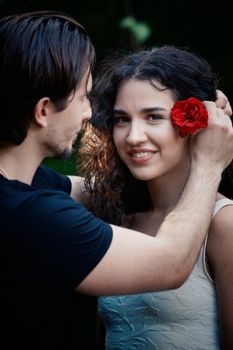 A couple shares a tender moment, adding a red flower to her hair outdoors.