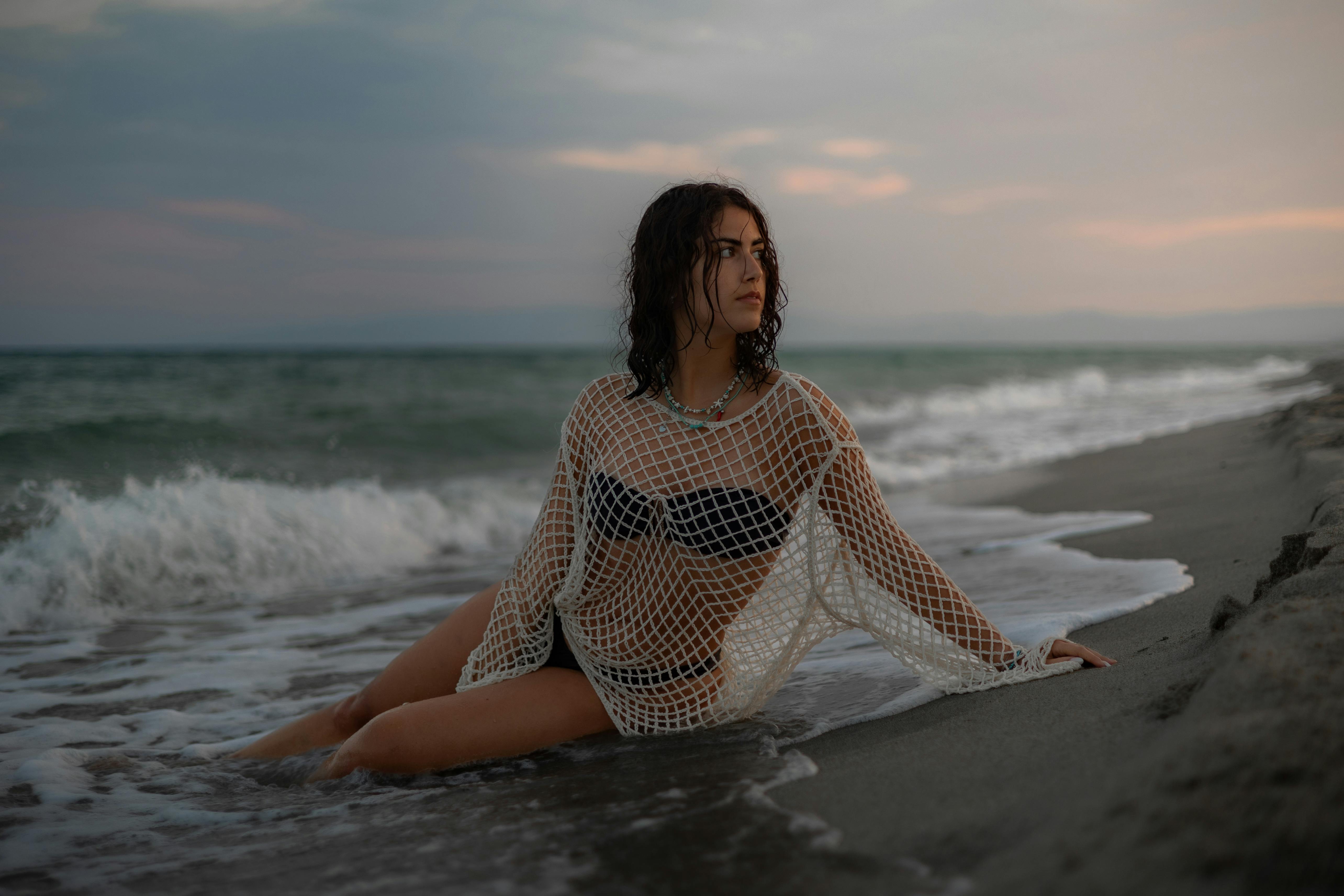 Woman in netted top sitting on Calabria beach at sunset, serene ocean vibes.