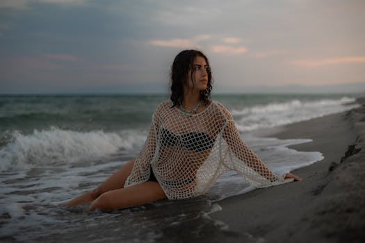Woman in netted top sitting on Calabria beach at sunset, serene ocean vibes.