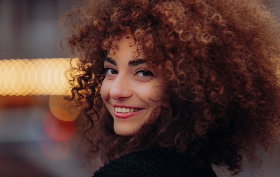 A striking portrait of a woman with curly hair smiling warmly, captured in soft natural light.
