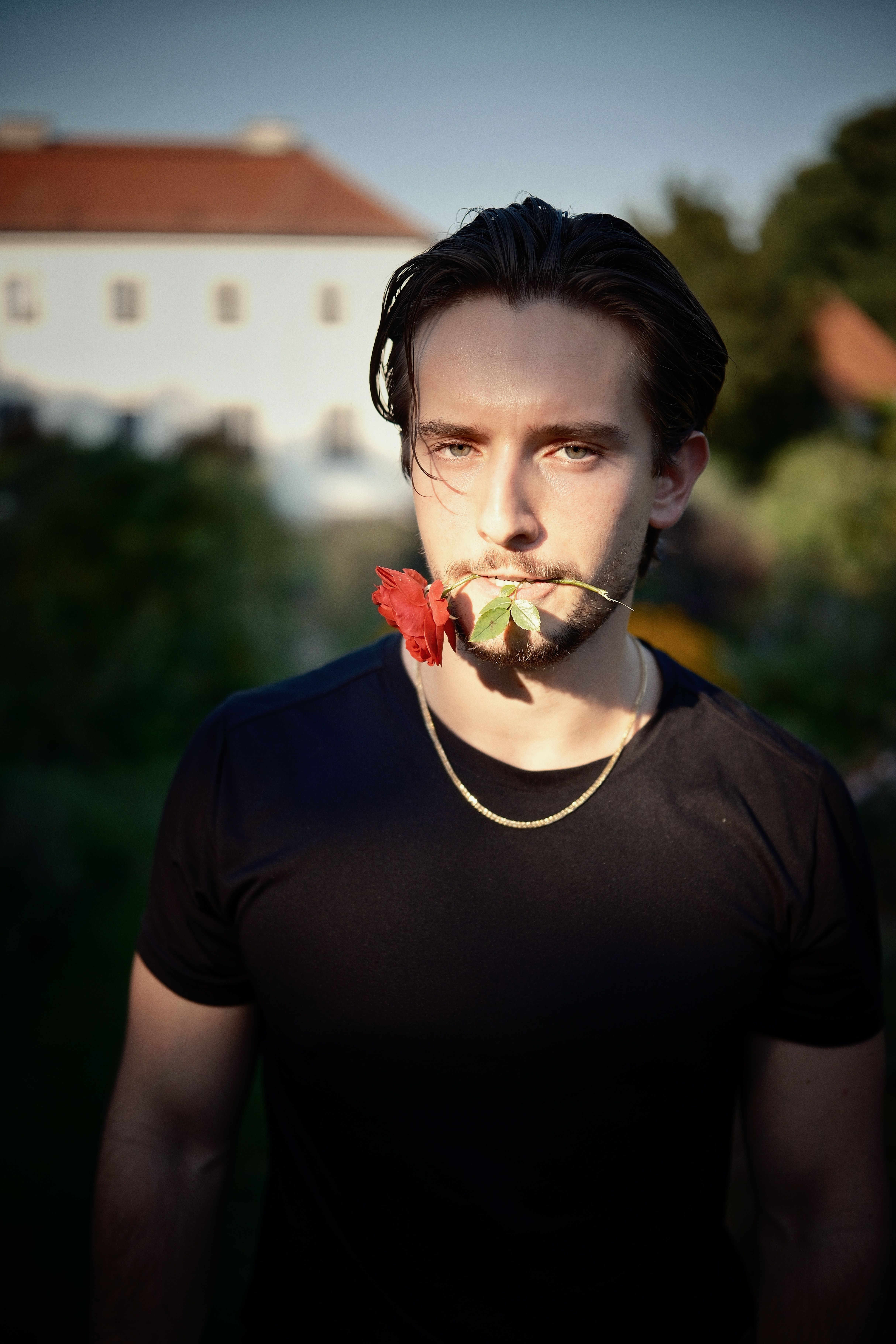 Young man with a rose in his mouth, standing outdoors in a garden during the day.