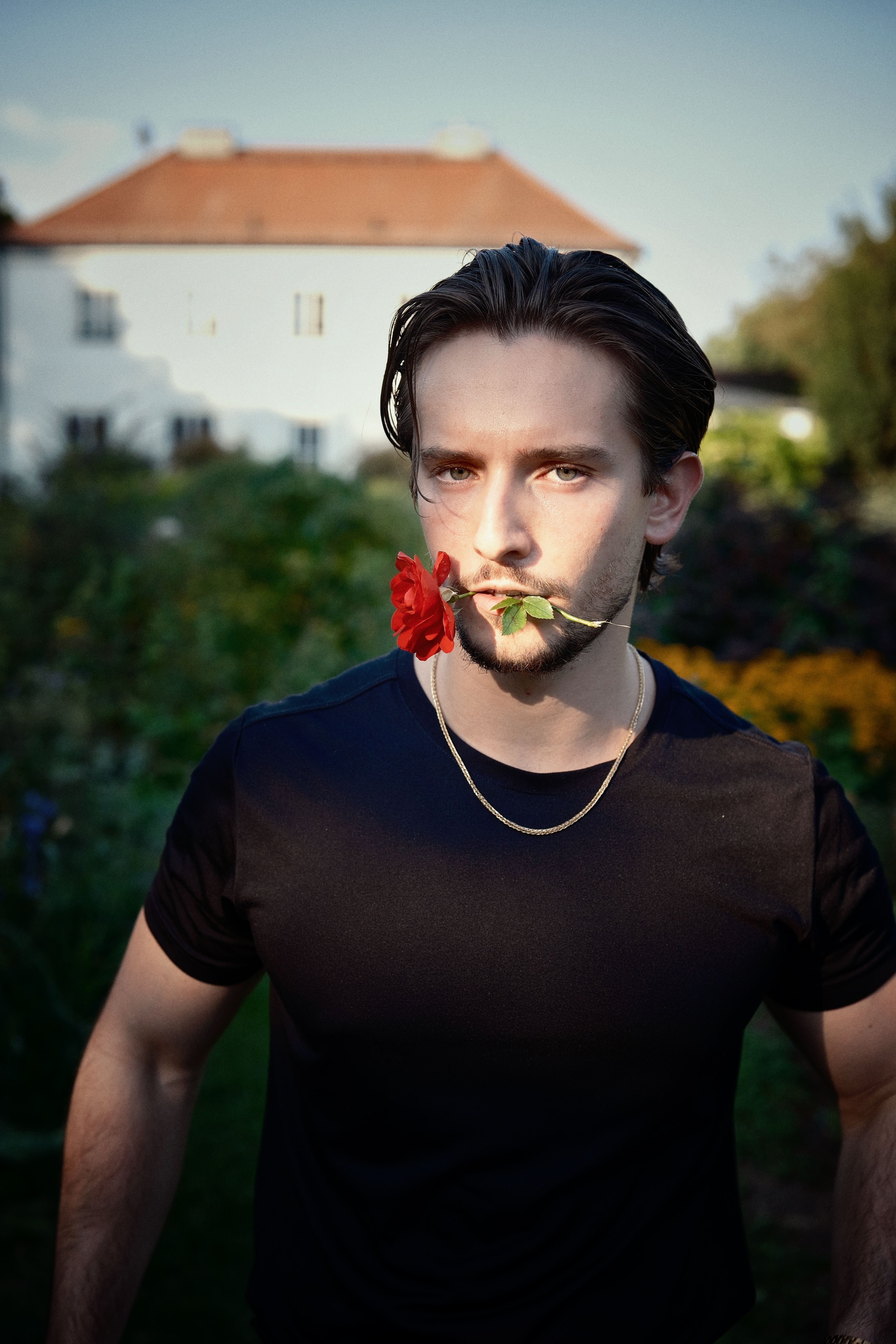 Portrait of a young man holding a red flower outdoors with sunlight and a building in the background.