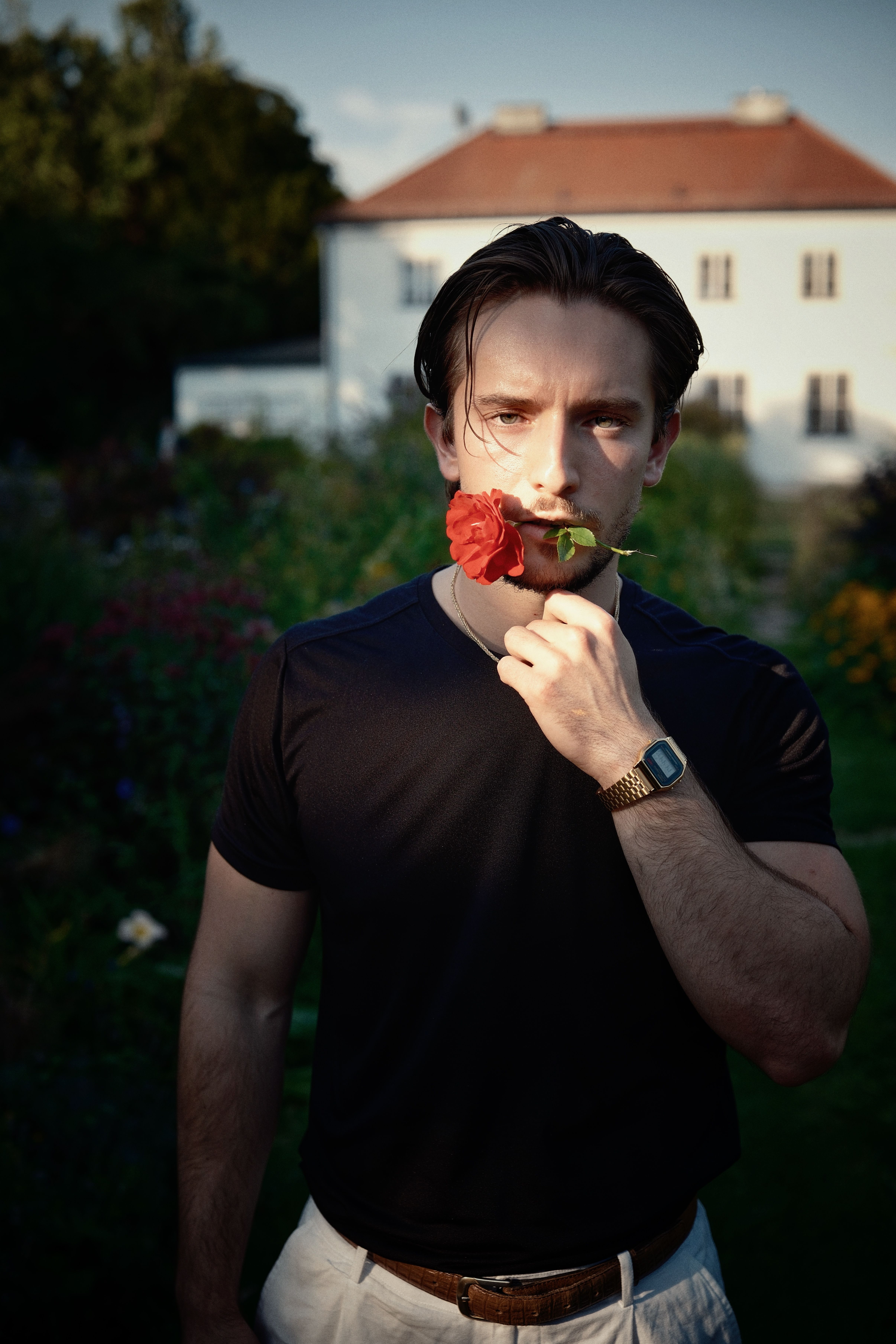 A man in a garden holds a red rose near his face, with a house in the background.