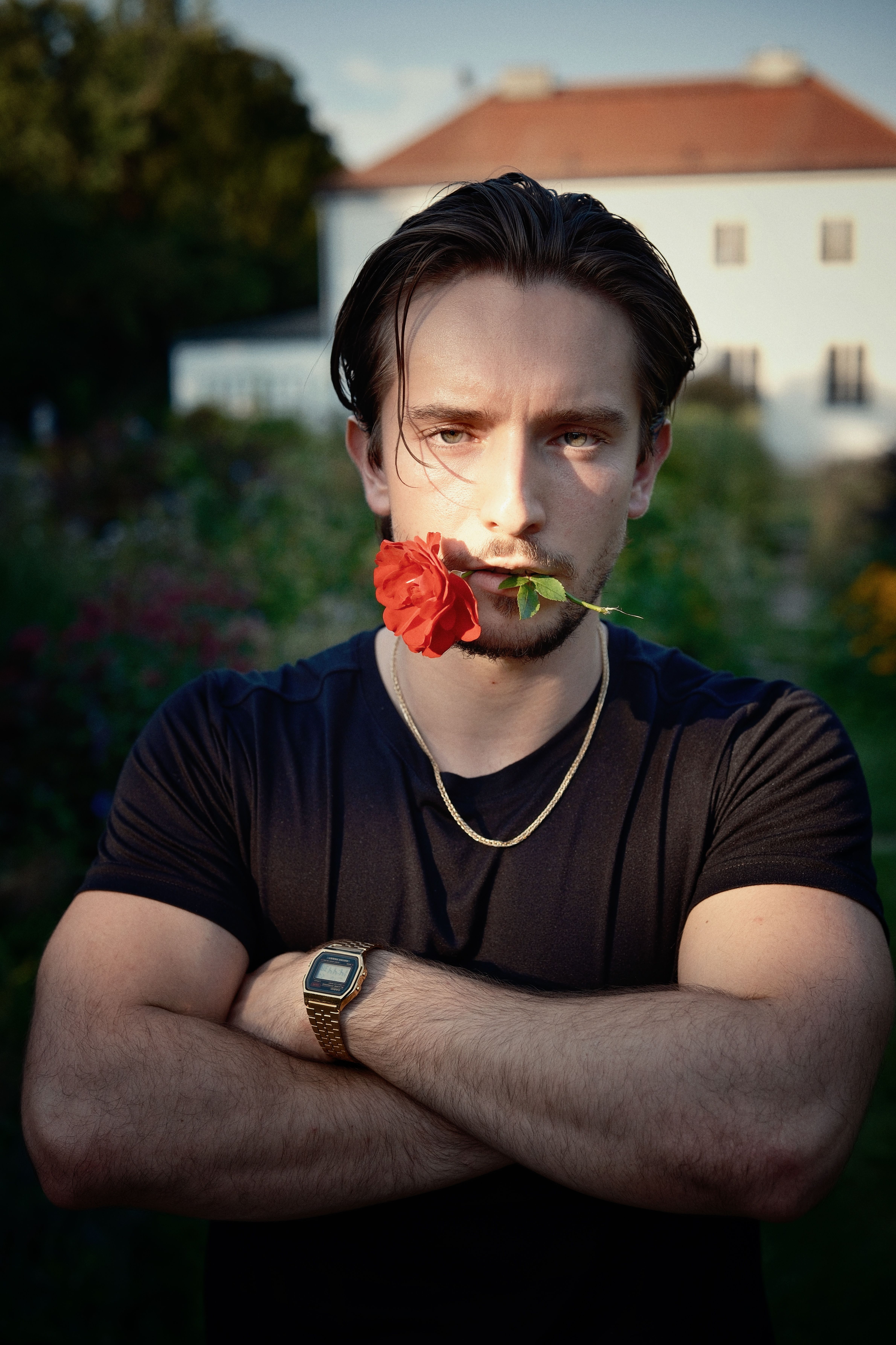A man with rose in mouth stands confidently in a garden, framed by a villa.