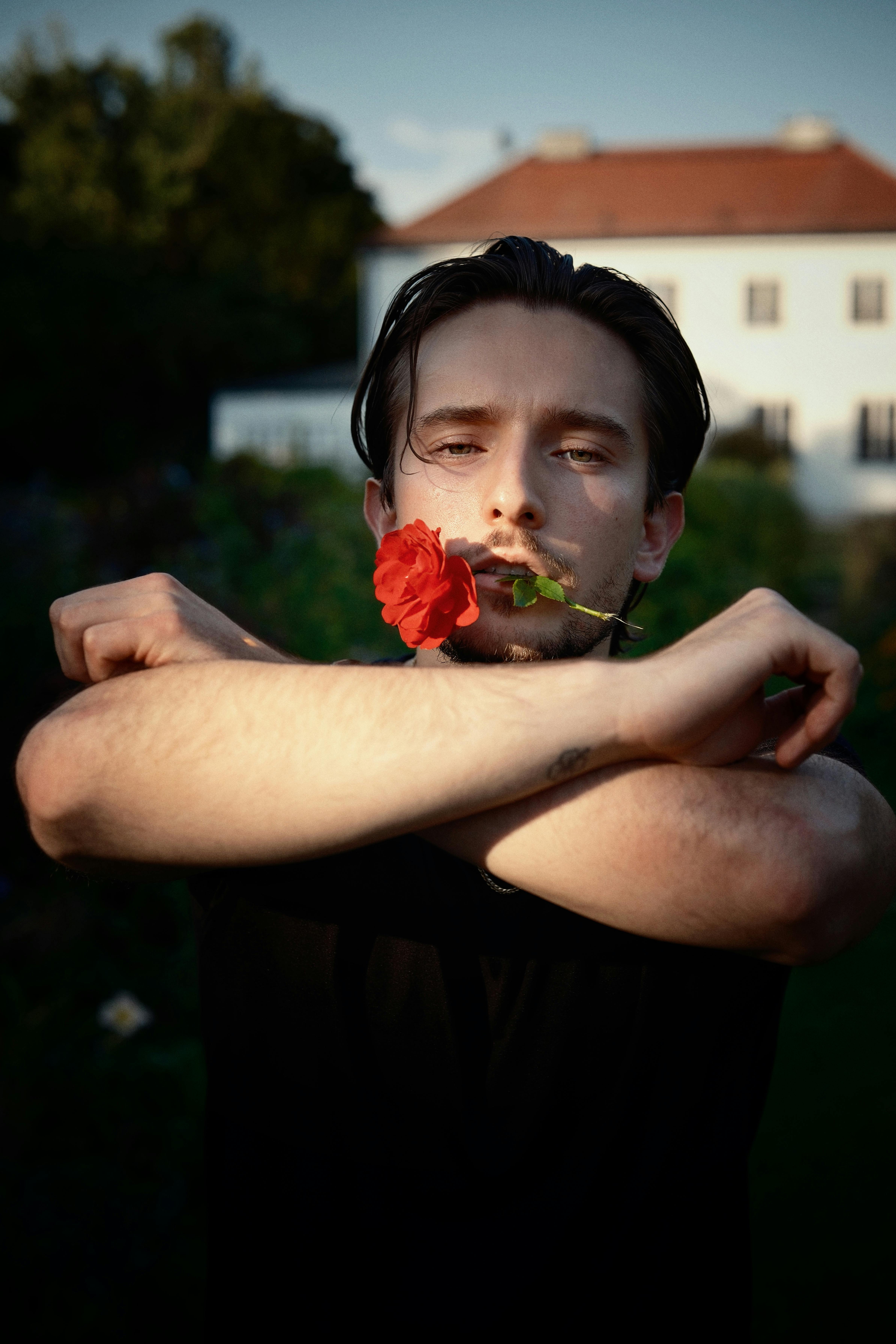 Portrait of a young man holding a red rose outdoors against a blurry building background.