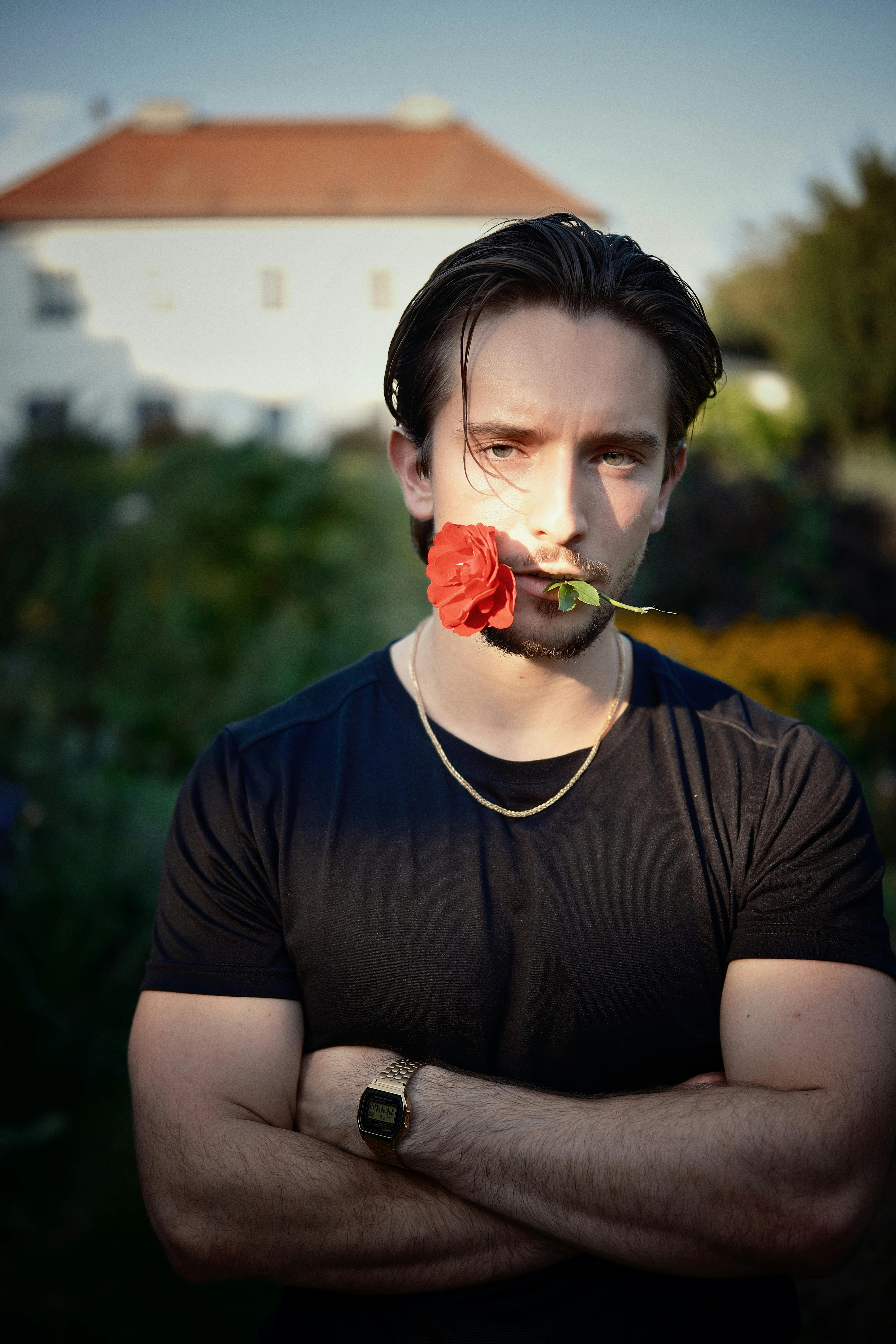 A man holding a rose in his mouth, standing outdoors with a garden background.