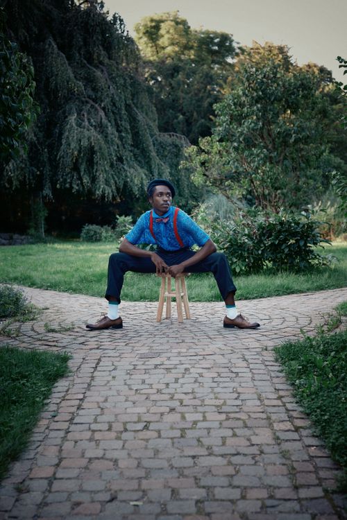 Free Young man with red suspenders sits on a stool in a lush park setting. Stock Photo