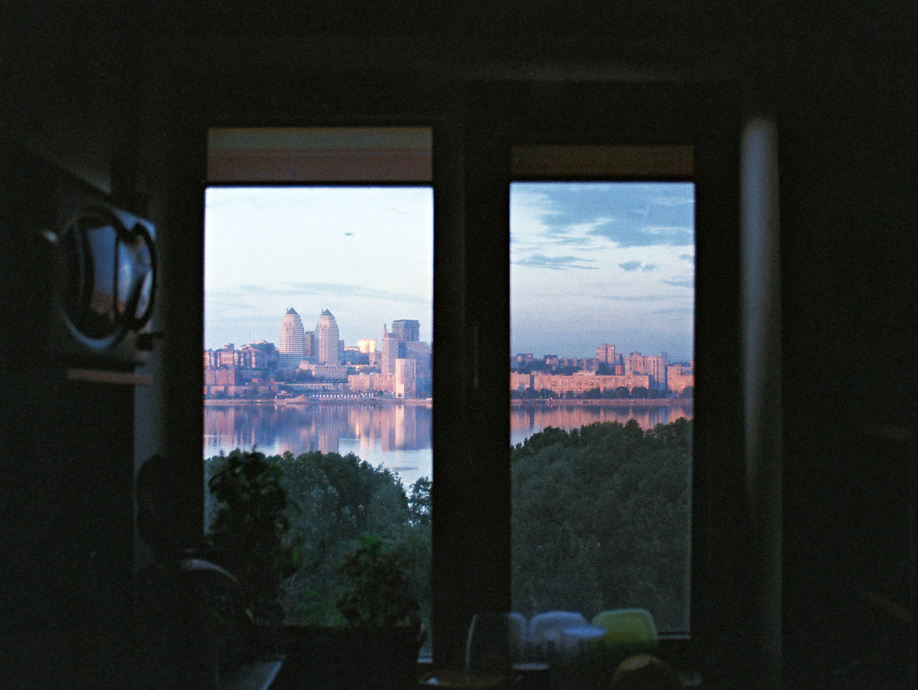 A serene view of Dnipro's skyline at dawn reflected in the river, captured through a window.