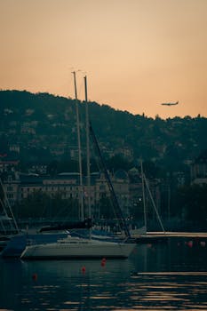 Calm waters and sailboats at sunset with an airplane in the sky, creating a serene cityscape.