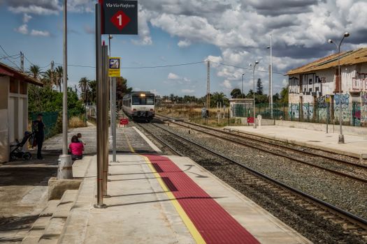 Train arrives at Torrellano station, Spain as people wait on a sunny day with clouds overhead.