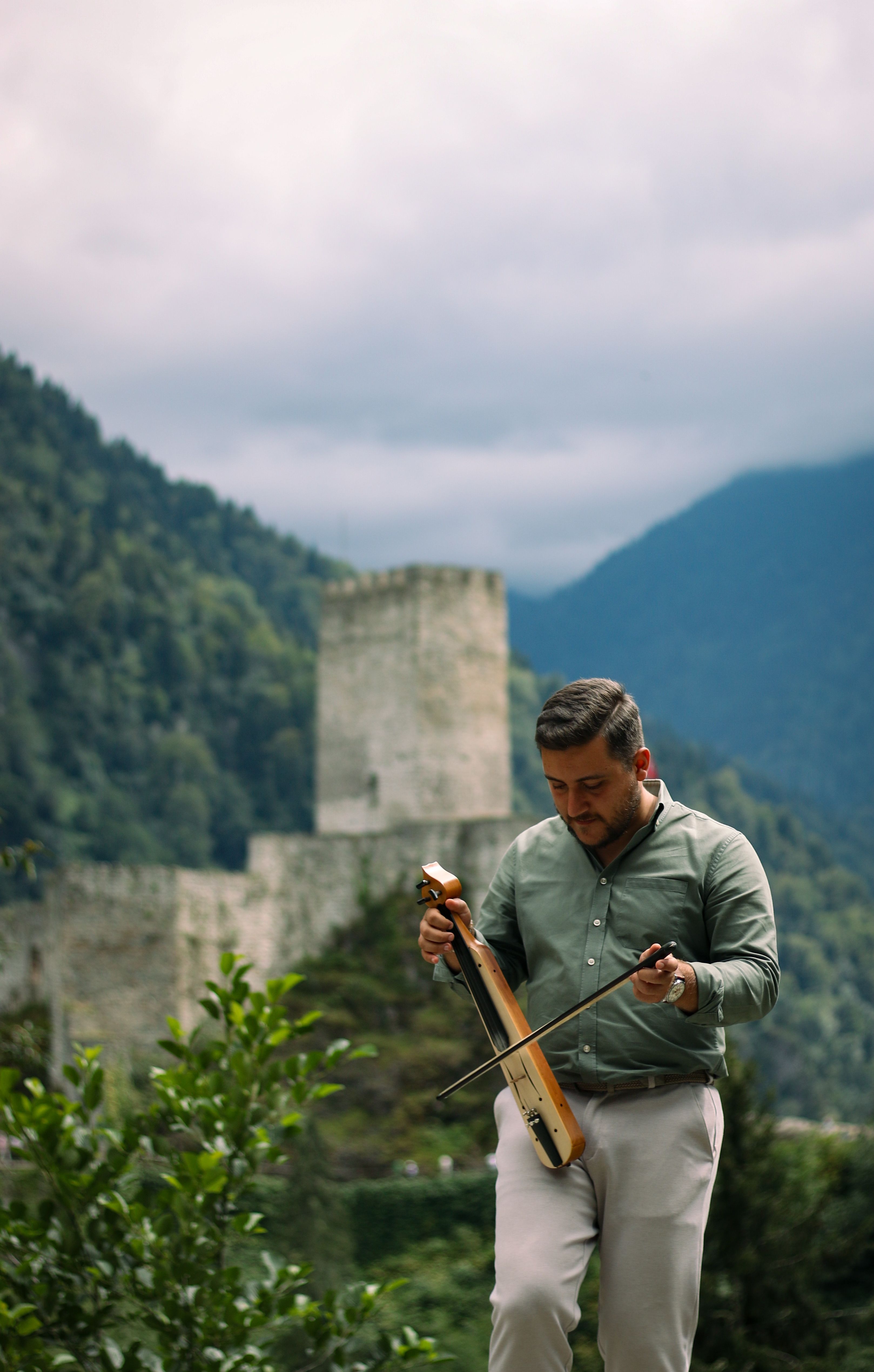 Man playing kamancheh near historic Zilkale Castle in Rize, Türkiye surrounded by lush greenery.