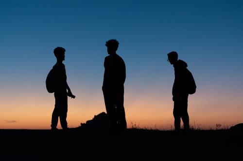 Free Three teenagers silhouetted against a vibrant sunset sky, standing outdoors with backpacks. Stock Photo