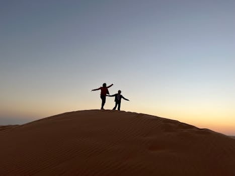 Two silhouetted figures stand atop a desert dune during twilight, capturing a peaceful moment.