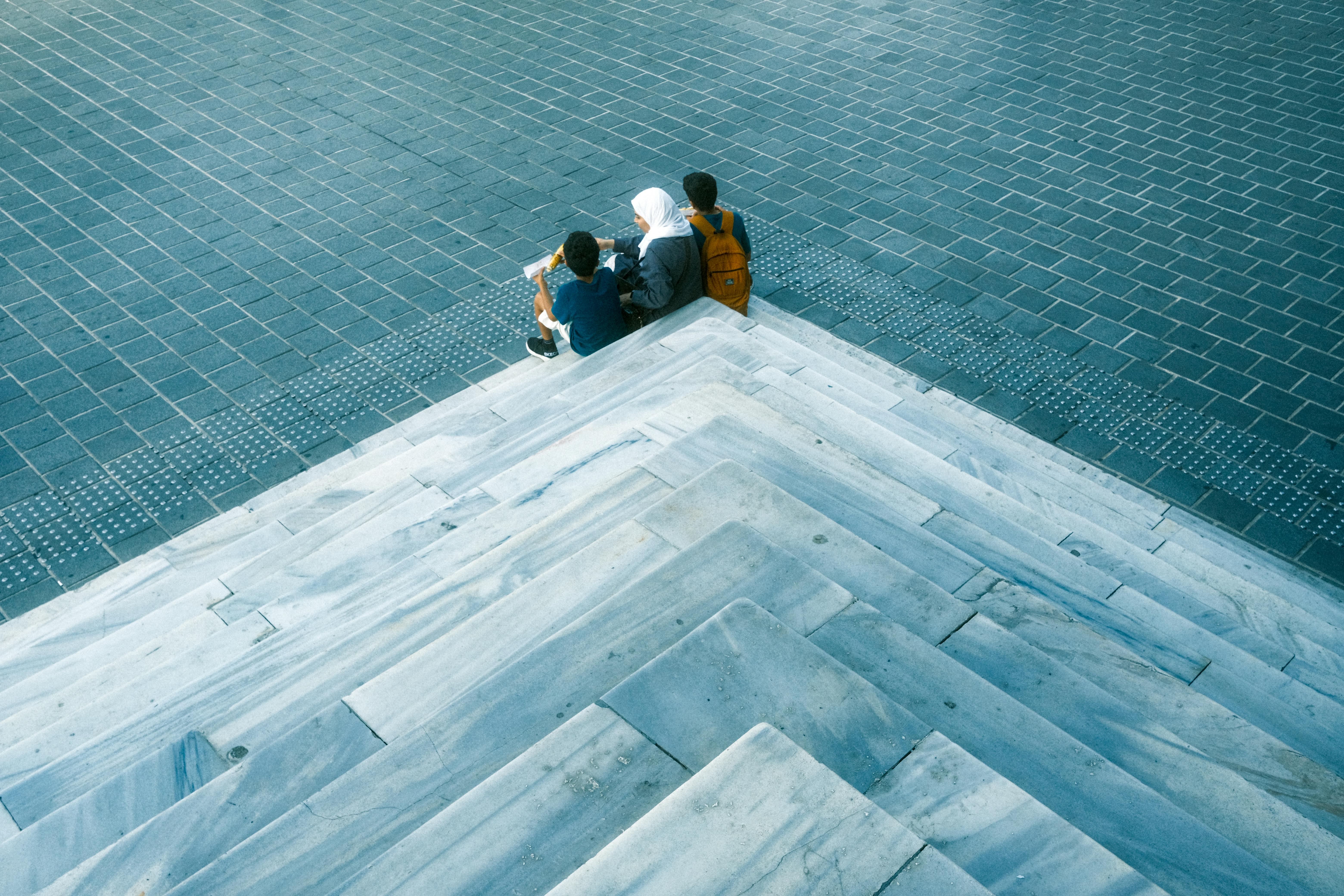 Kostenlos Eine Gruppe von Menschen sitzt auf Marmorstufen, im Hintergrund ist ein städtisches Pflaster zu sehen. Stock-Foto
