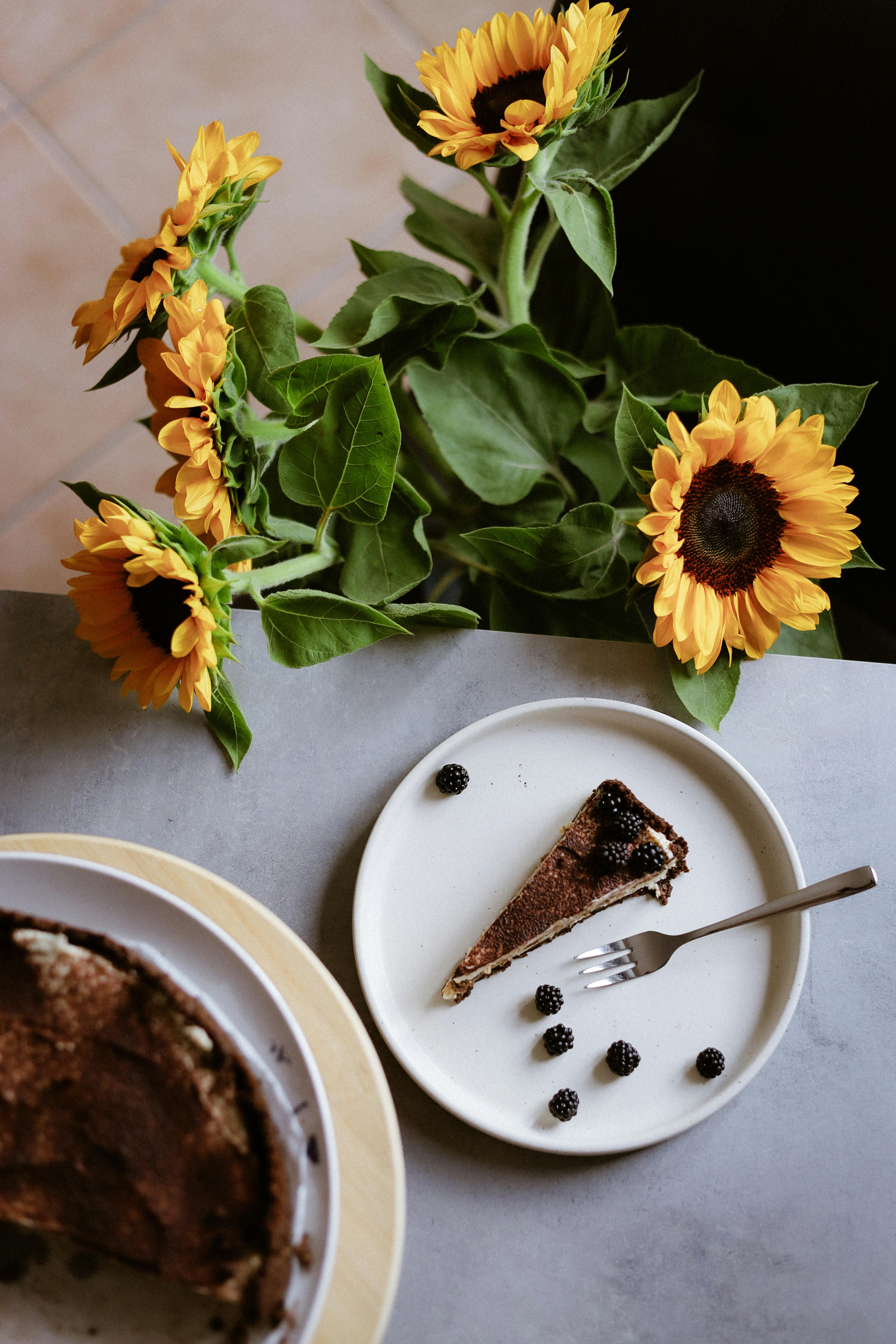 Top view of chocolate cake slice with blackberries beside vibrant sunflowers.