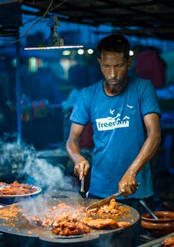 A street vendor prepares spicy food with intense focus at a bustling night market in Dhaka, Bangladesh.