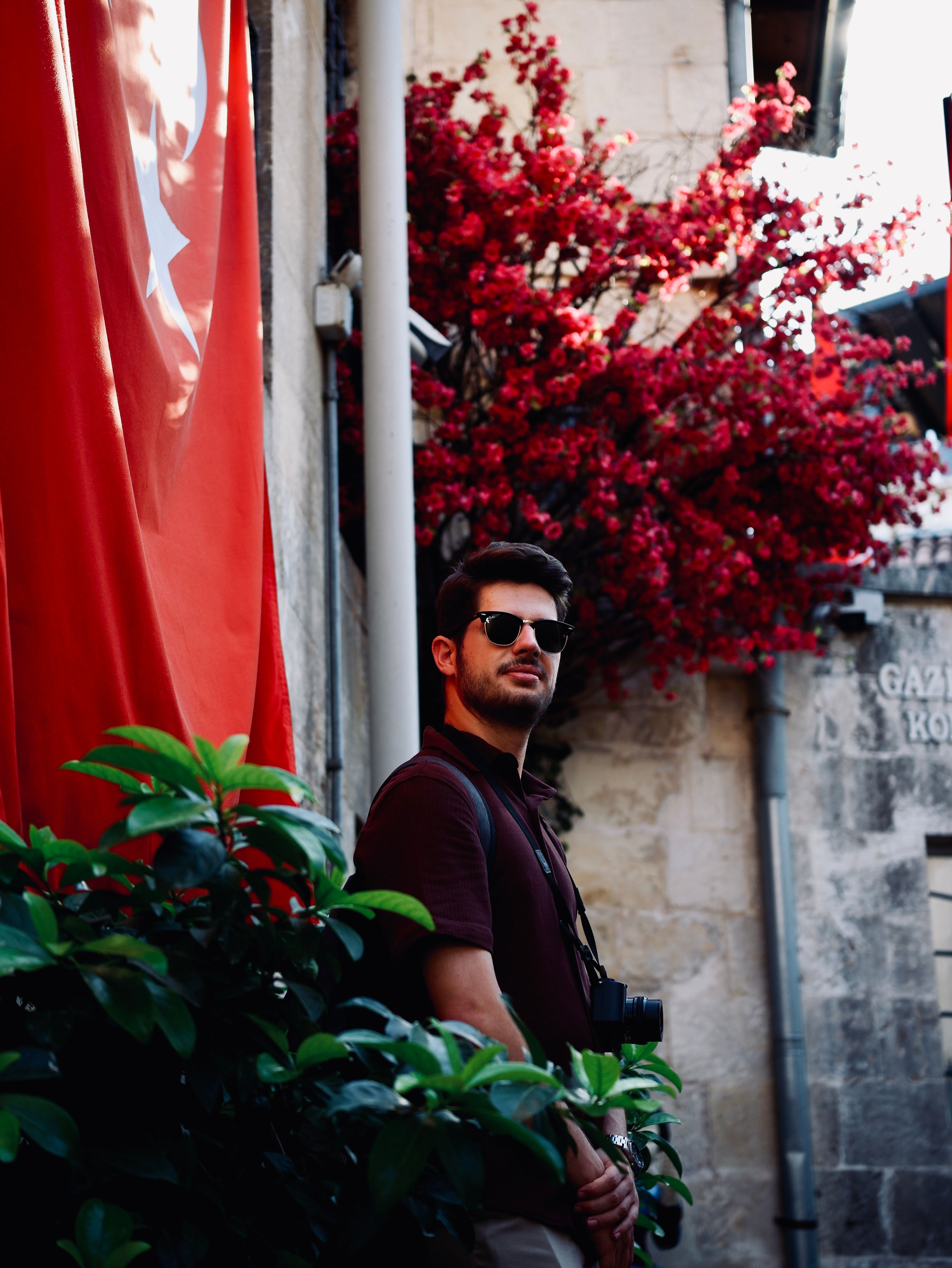 Fashionable man in sunglasses poses in scenic alley with red flowers and flag backdrop.