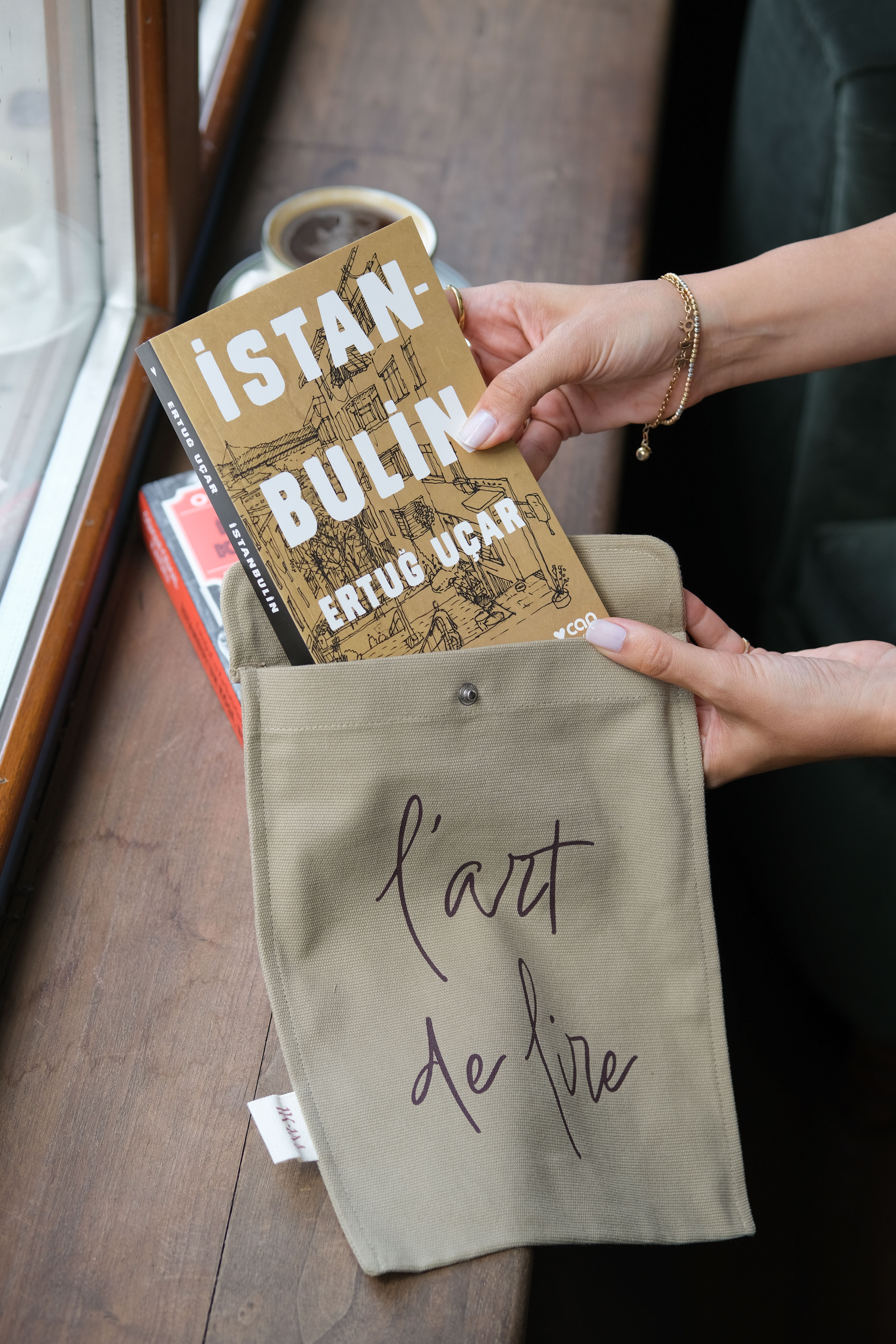 Hands place an 'İstanbul'un' book into a canvas bag near window.