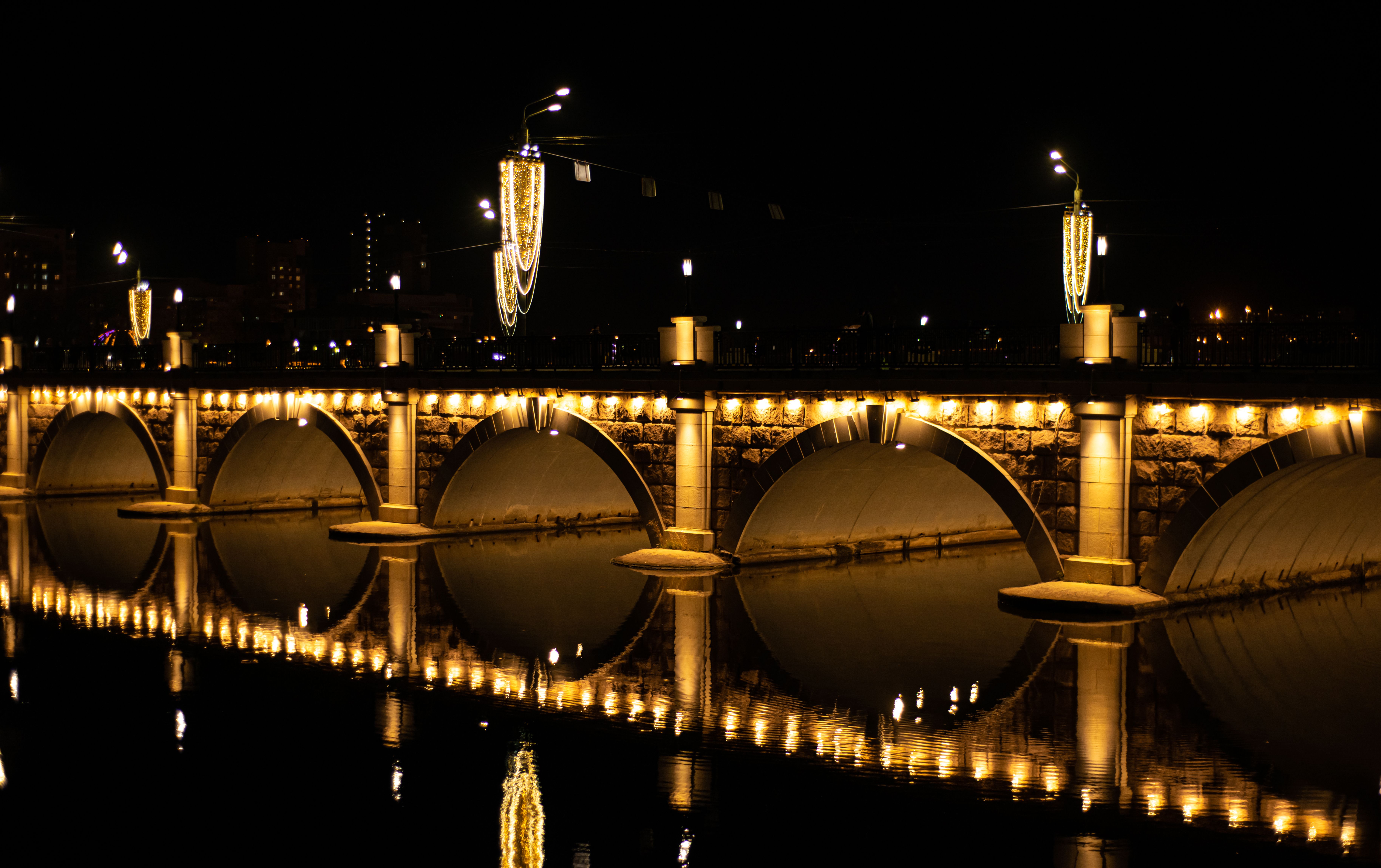Night view of a lit stone bridge with reflections on water, creating a symmetrical pattern.
