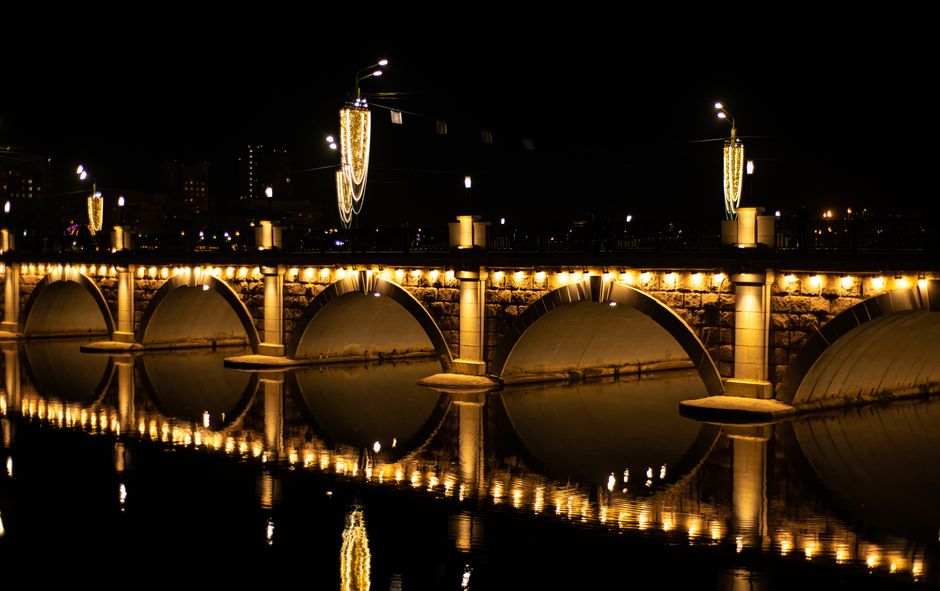 Night view of a lit stone bridge with reflections on water, creating a symmetrical pattern.