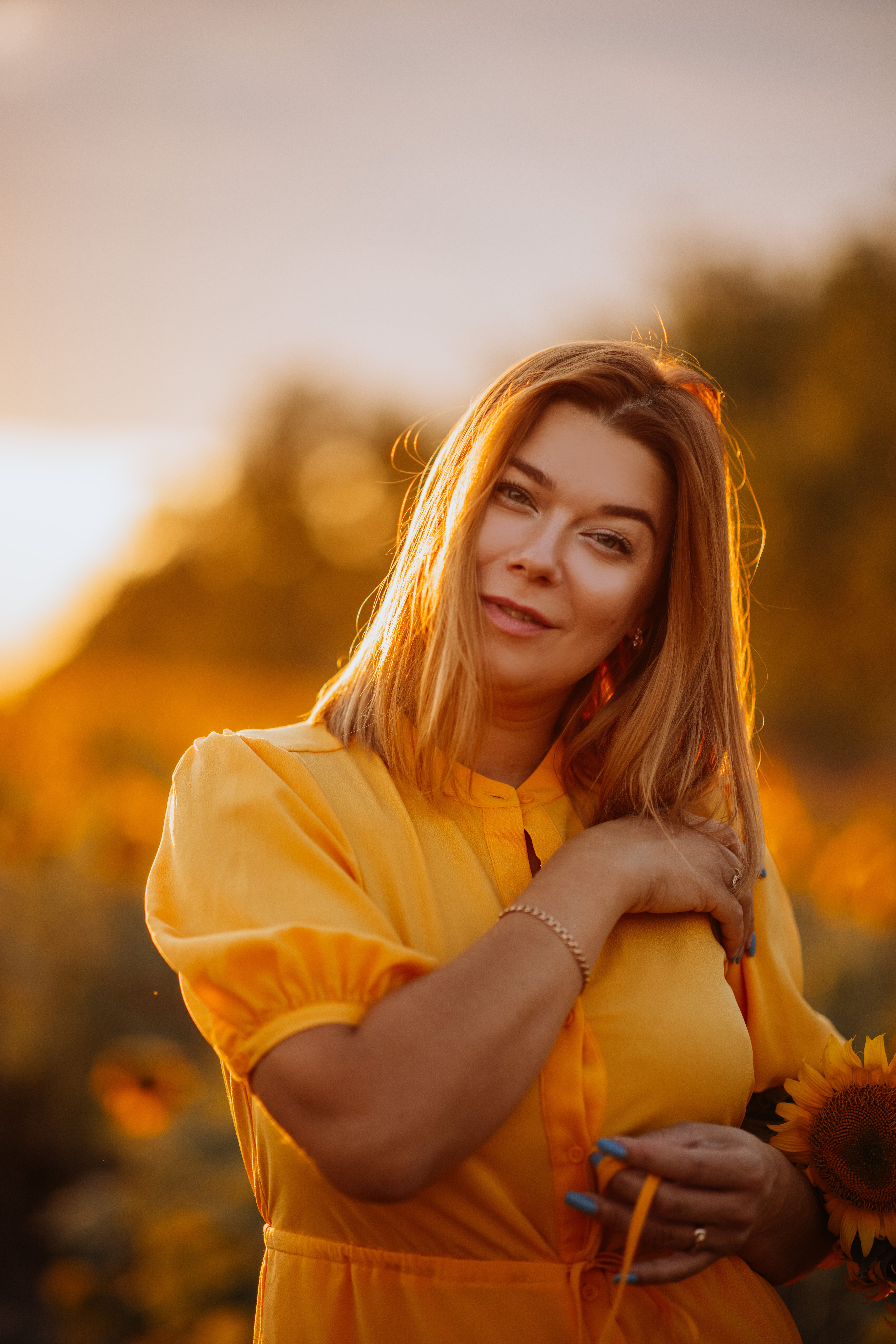 A woman in a yellow dress smiles while holding a sunflower in a field during sunset.