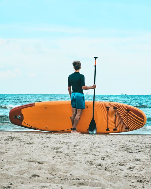 Free Man standing on sandy beach with paddleboard, gazing at the ocean. Stock Photo