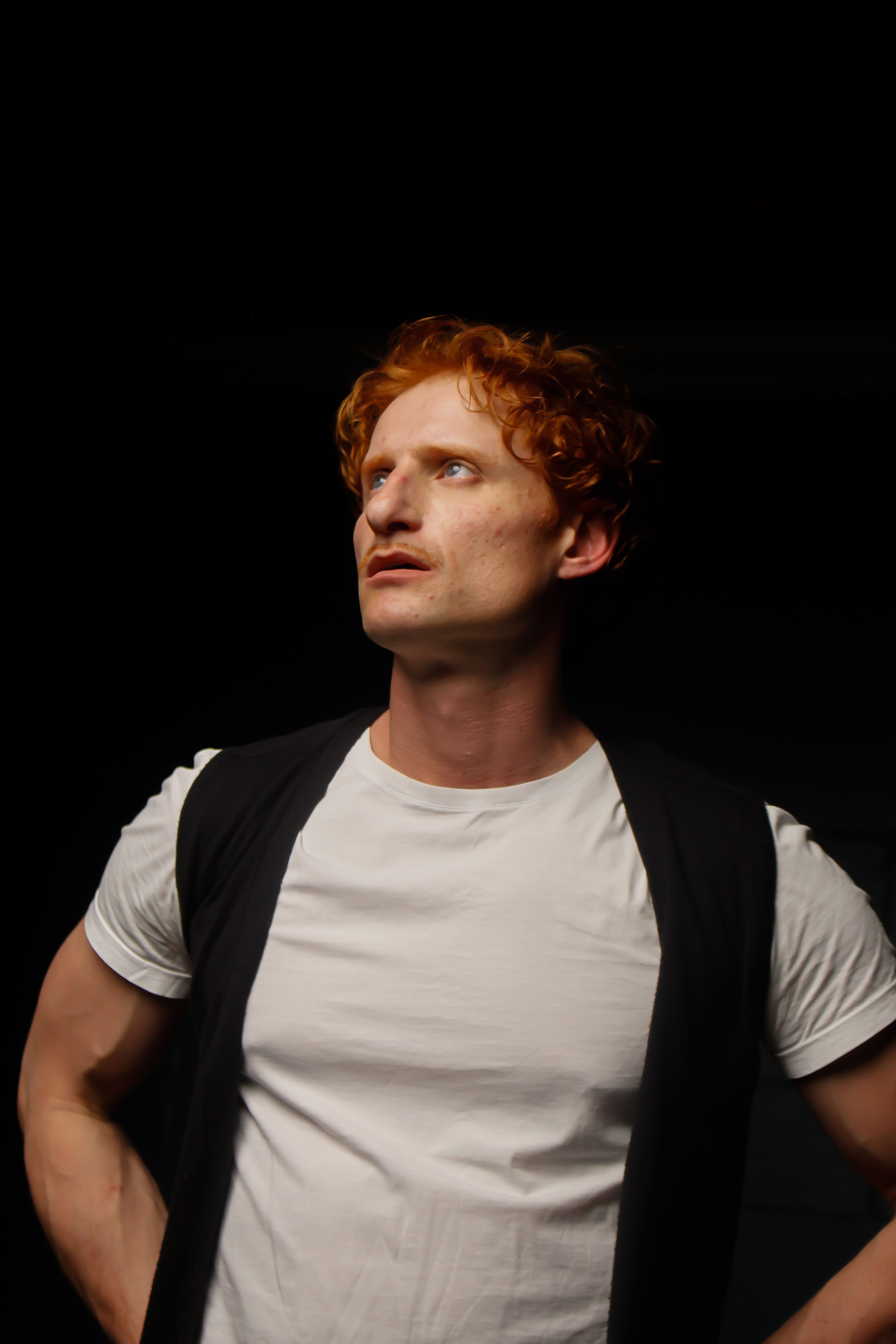 A man with curly red hair looks pensively upward, captured in dramatic lighting.