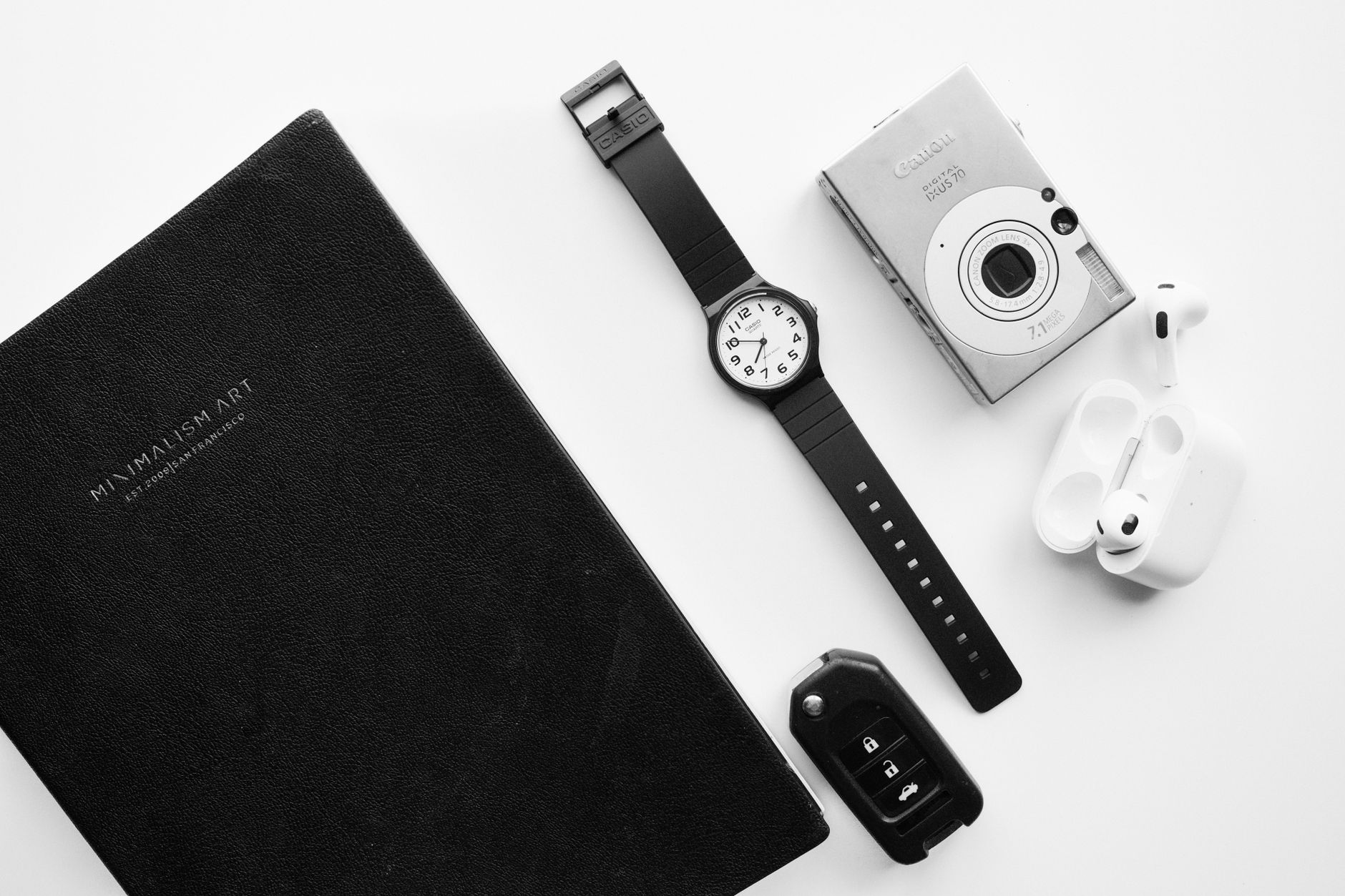 A black and white flat lay featuring a notebook, wristwatch, digital camera, earphones, and car key