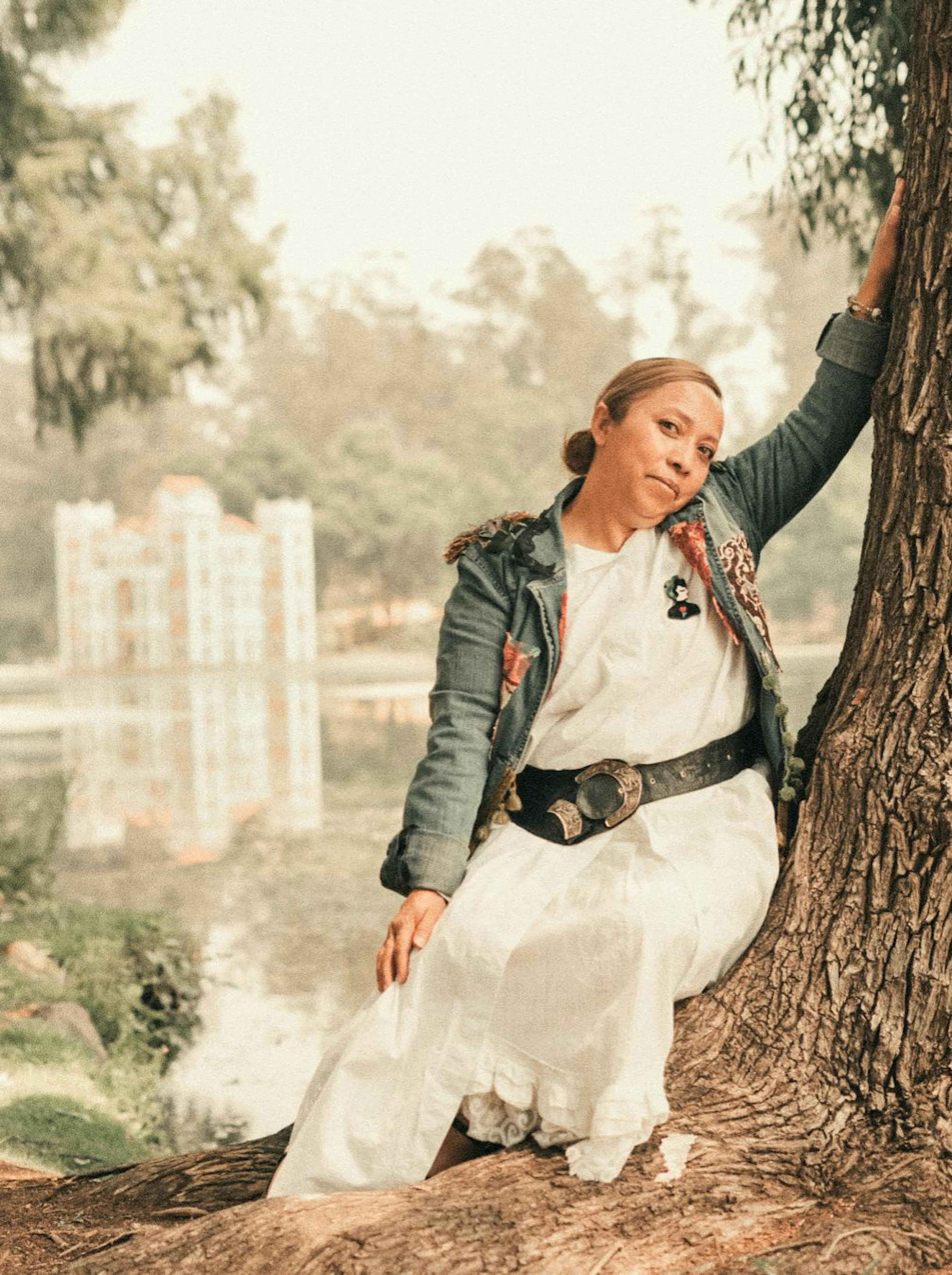 Woman in casual attire sitting by a tree near a serene lake with trees.