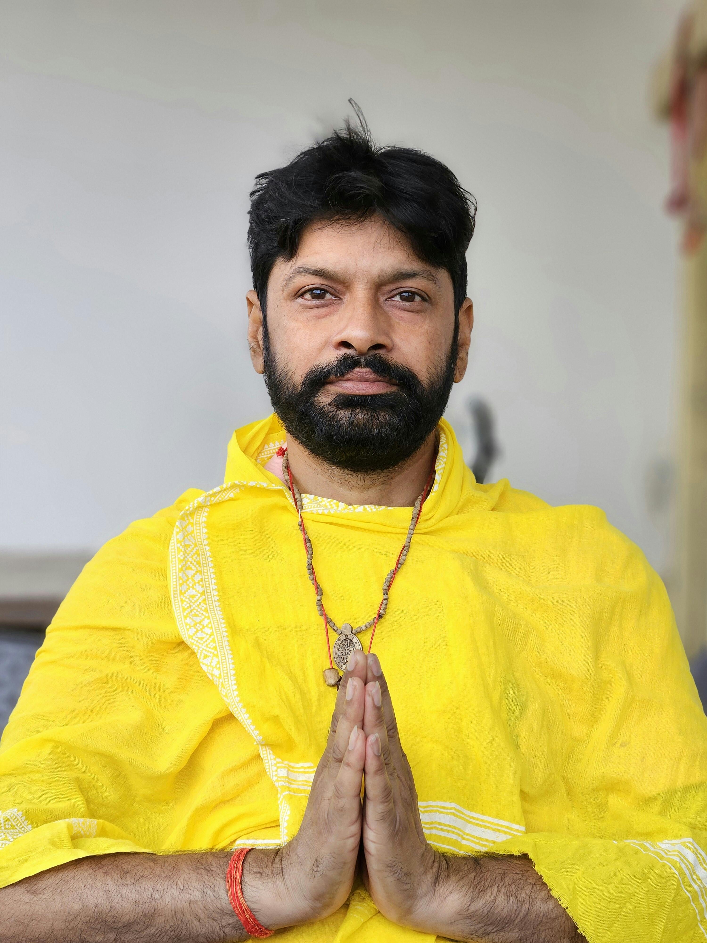 Man in yellow traditional attire meditating with namaste gesture indoors.