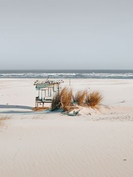 A lonely beach hut surrounded by sand dunes and ocean waves under a clear sky.