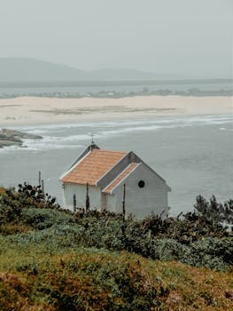A small white chapel overlooking the ocean, surrounded by lush greenery and hills.