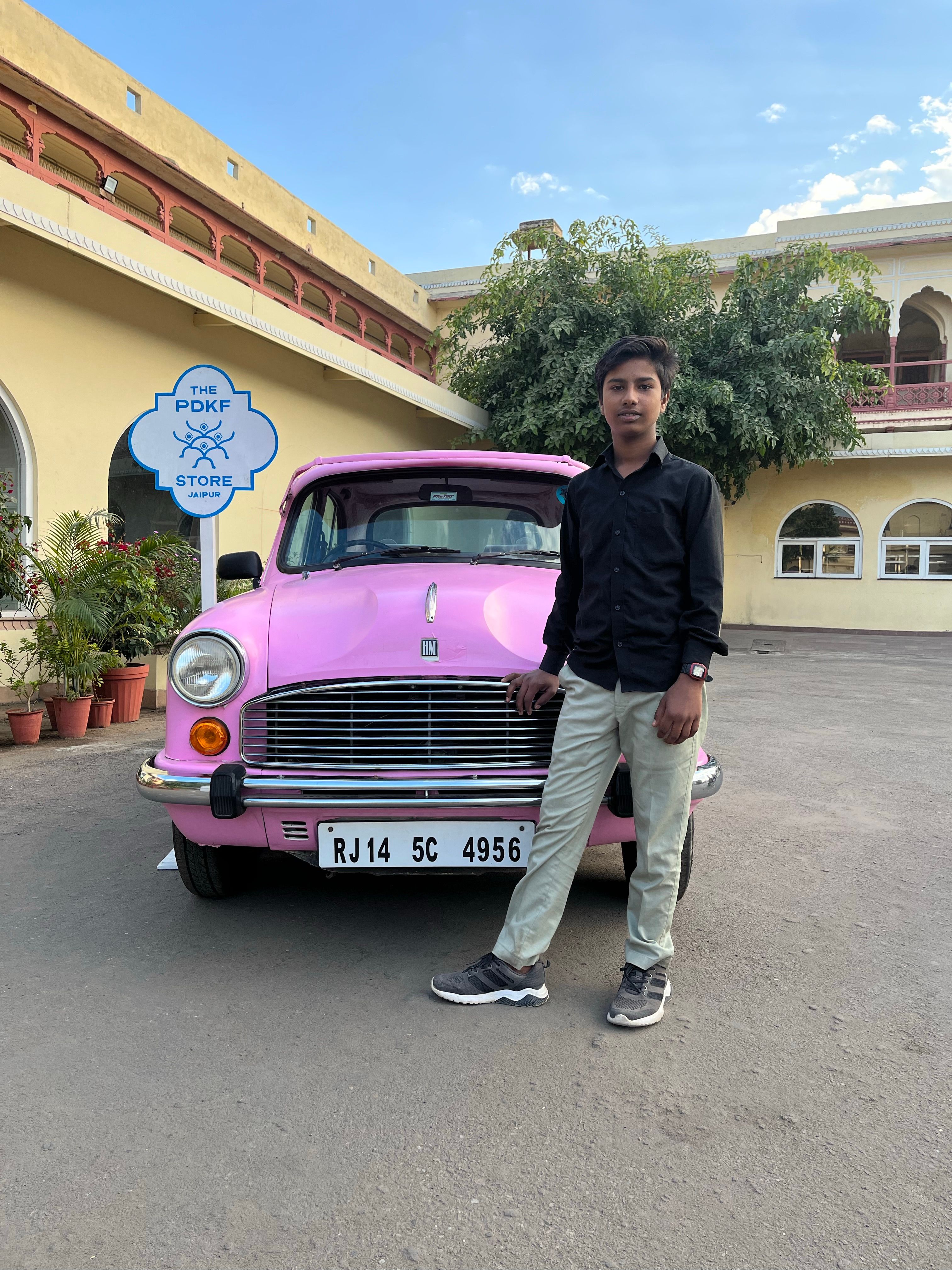 Young man standing by a classic pink car in front of a heritage building, showcasing style and vintage charm.