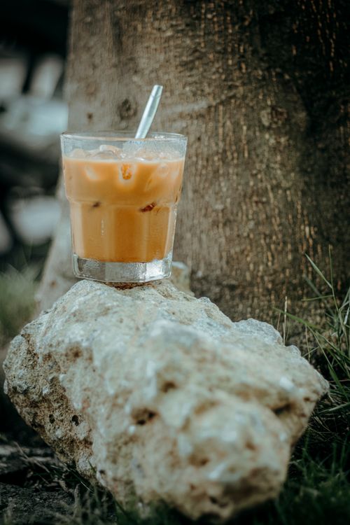 Free A glass of iced coffee with a straw placed on a rock by a tree outdoors. Stock Photo