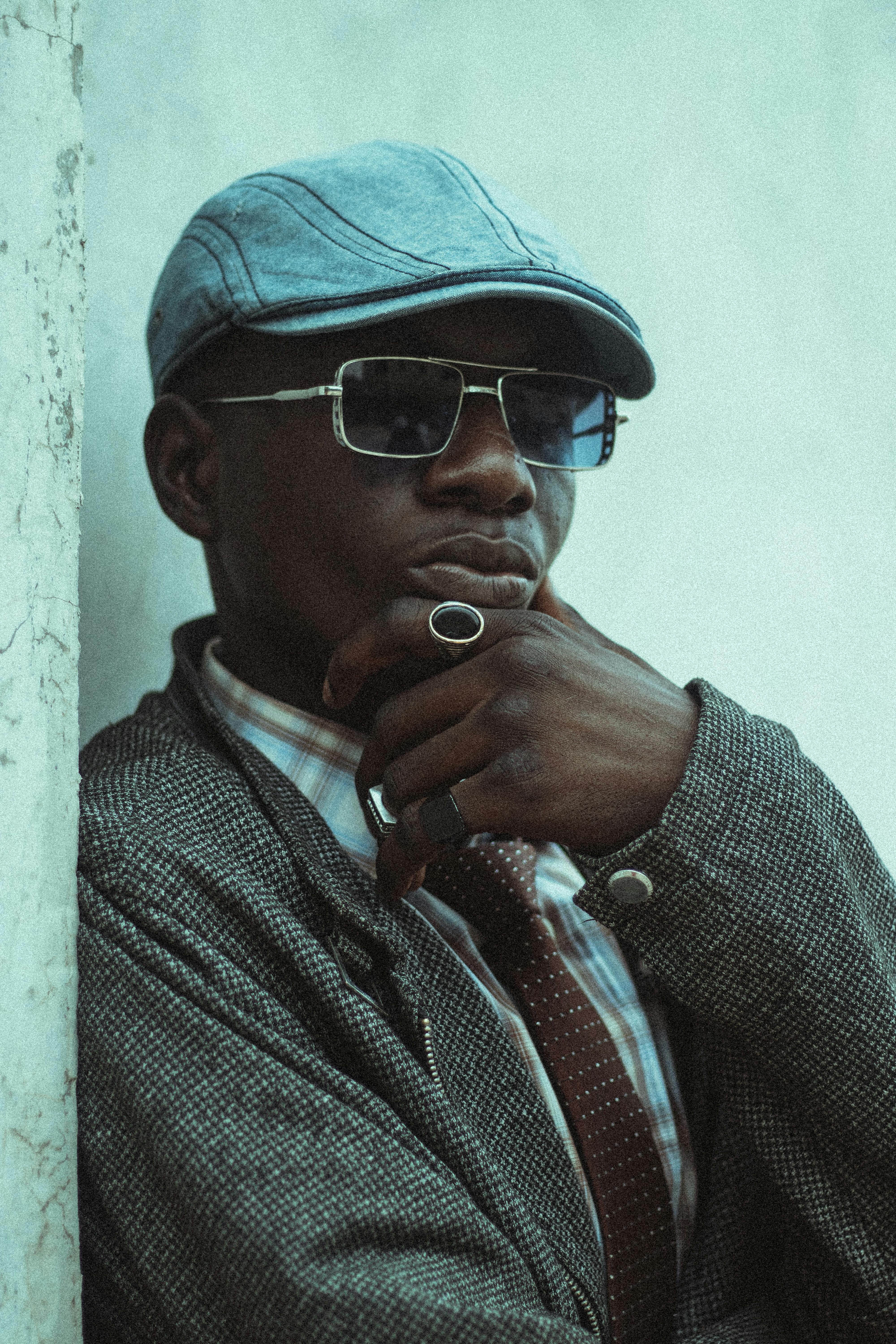 A fashionable man wearing sunglasses and a hat poses with confidence against a rustic wall.