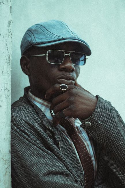 A fashionable man wearing sunglasses and a hat poses with confidence against a rustic wall.