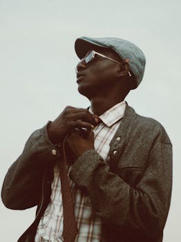 Man in a cap adjusting his tie, wearing sunglasses and a jacket outdoors.