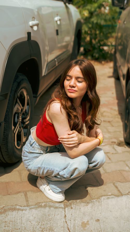 Free A stylish young woman squats between cars on a sunny urban street, exuding confidence. Stock Photo