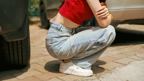 Free Side view of a person squatting outdoors, showcasing casual street fashion with a red top and jeans. Stock Photo