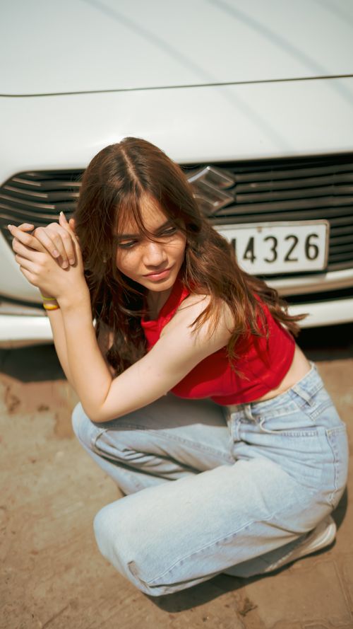 Free Casual portrait of young woman in red top and jeans squatting near car, outdoors, daytime. Stock Photo